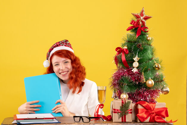 圣诞节正面图：女医生坐在桌子后面 拿着黄色背景上的文件 还有圣诞树和礼品盒坐着抱着生活