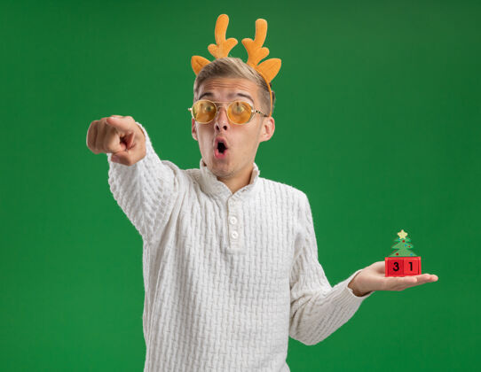 指向给人印象深刻的年轻帅哥戴着驯鹿鹿角头带戴着眼镜拿着圣诞树玩具看着日期指着隔离在绿色墙上的一面头带穿着印象深刻