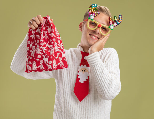 保管微笑着的年轻帅哥戴着圣诞新奇眼镜打着圣诞老人的领带拿着圣诞袋看着相机手放在橄榄绿背景上孤立的脸上帅哥领带抱着