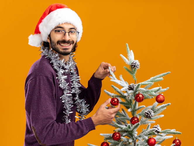 就在附近微笑的年轻帅哥站在圣诞树旁 戴着圣诞帽 脖子上戴着花环 橙色的背景孤立着帅气年轻花环