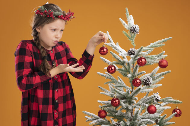 圣诞节小女孩戴着圣诞花圈 穿着格子裙装饰圣诞树 站在橙色的背景下 显得困惑和焦虑困惑焦虑橙色