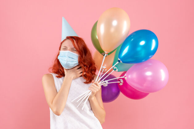 年轻正面图是粉红色墙上戴着消毒面具手持彩色气球的年轻女子生日多彩丝带