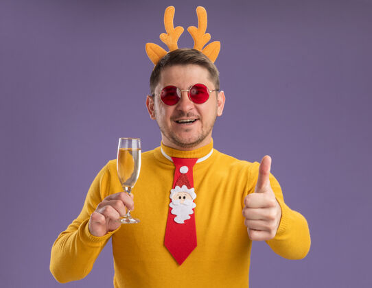 眼镜穿着黄色高领毛衣 戴着红色眼镜 戴着有趣的红色领带和鹿角边 手持香槟酒杯 竖起大拇指站在紫色背景上的快乐年轻人角轮辋香槟酒