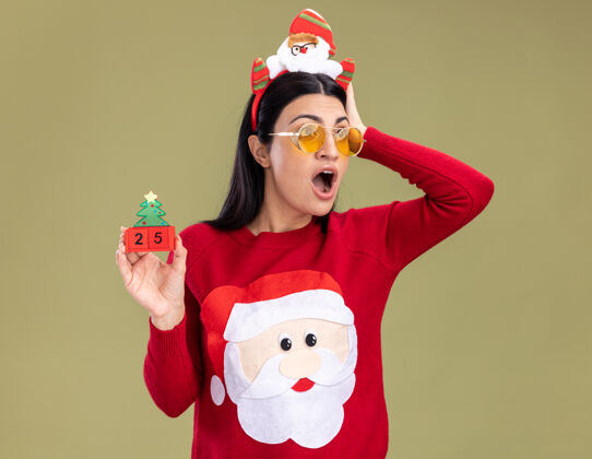 穿着关心的年轻白人女孩戴着圣诞老人的头带和带眼镜的毛衣拿着圣诞树玩具和日期手放在头上看着橄榄绿墙上孤立的一面日期保管关心