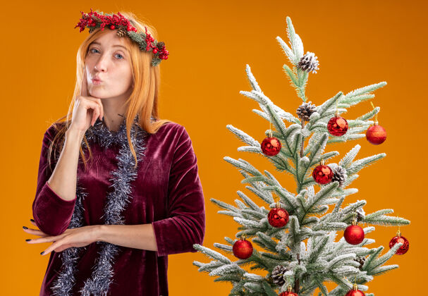花环令人印象深刻的噘嘴年轻漂亮的女孩站在圣诞树附近穿着红色的裙子和花环与花环的脖子上把手指放在脸颊隔离在橙色的墙上站着花环圣诞节