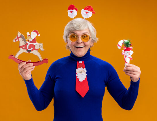 头带令人印象深刻的老太婆在太阳眼镜与圣诞老人头带和圣诞老人领带举行圣诞老人的摇马装饰和糖果手杖孤立的橙色背景与复制空间眼镜老人摇摆