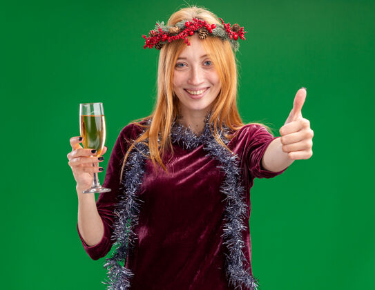 抱着微笑着的年轻漂亮的女孩穿着红色的裙子 脖子上戴着花环和花环 手里拿着一杯香槟 在绿色的背景上孤立地竖起大拇指花环香槟微笑