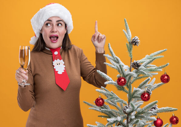 附近给人印象深刻的年轻漂亮女孩戴着圣诞帽打着领带站在圣诞树旁拿着一杯香槟点在橙色的墙上孤立起来领带帽子穿着