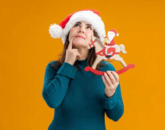 橙色体贴的成年白人妇女戴着圣诞老人帽 把手指放在下巴上 把圣诞老人抱在摇马装饰上 在橙色背景上孤立地向上看 还有复制空间帽子手指持有