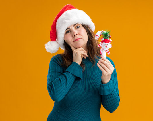橙色困惑的成年白人妇女戴着圣诞帽 把手指放在下巴上 拿着橙色背景上的糖果手杖 还有复制空间困惑手杖帽子