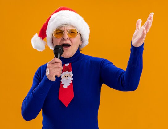 假装戴着太阳眼镜 戴着圣诞帽 系着圣诞领带的快乐的老妇人拿着麦克风 假装在橙色背景上孤立地唱歌 还有复制空间眼镜圣诞老人快乐