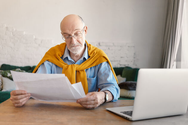 办公室戴着长方形眼镜的雅致胡须老人 手里拿着纸 在家里在线计算家庭财务 在客厅内部使用电子便携设备人成人老年人