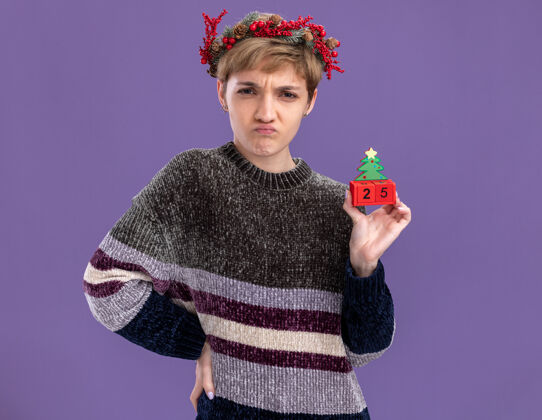 皱眉皱眉的年轻漂亮女孩戴着圣诞花环手持圣诞树玩具手放在腰上日期隔离在紫色的墙上头圣诞节日期
