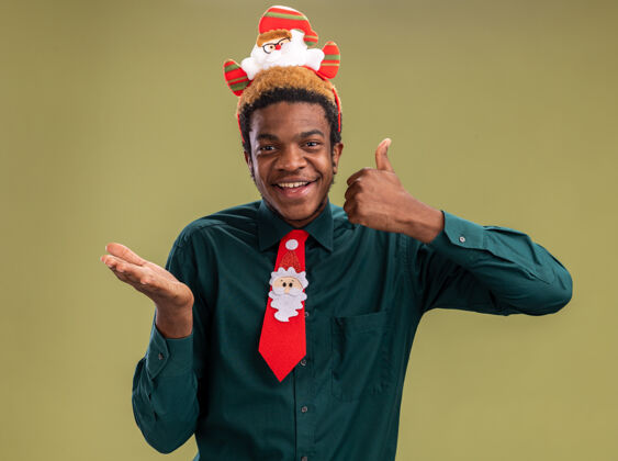 非洲快乐的非洲裔美国人 戴着有趣的圣诞老人戒指 打着红色领带 站在绿色背景上 看着相机上下竖起大拇指圣诞老人美国人领带