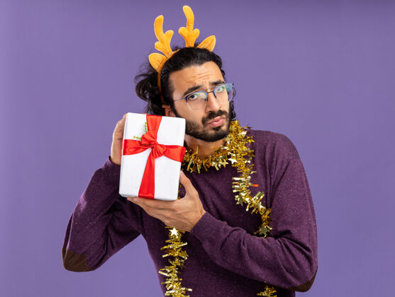 头发年轻帅哥戴着圣诞发箍 脖子上戴着花环 手里拿着蓝色背景的礼品盒小伙子年轻人盒子