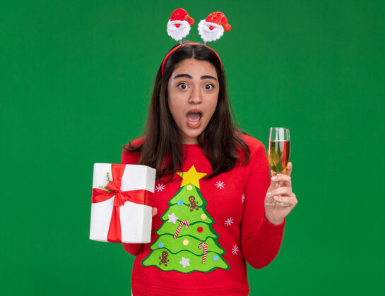 盒子焦虑的年轻白人女孩戴着圣诞老人的头带 手里拿着一杯香槟和一个圣诞礼盒 背景是绿色的 还有复印空间杯子圣诞节年轻