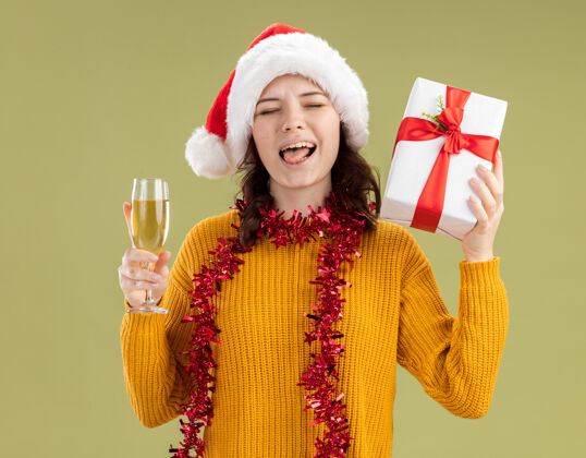 快乐快乐的年轻斯拉夫女孩戴着圣诞帽 脖子上戴着花环 伸出舌头 拿着一杯香槟和圣诞礼盒 橄榄绿的背景上有复制空间圣诞老人舌头花环