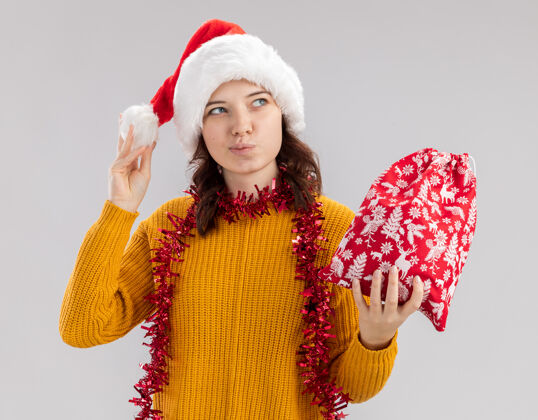 花环年轻的斯拉夫女孩戴着圣诞帽 脖子上戴着花环 手里拿着圣诞礼品袋 一边看着白色背景上孤立的一面 一边留着复制空间举行周围高兴