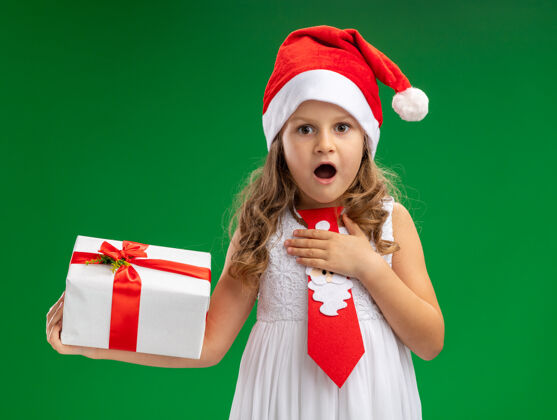 她自己惊讶的小女孩戴着圣诞帽打着领带拿着礼品盒把手放在自己的绿色背景隔离惊讶手领带