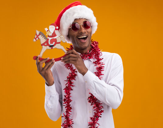 摇摆年轻的美国黑人男子戴着圣诞帽 戴着眼镜 脖子上戴着金箔花环 抱着摇摇木马上的圣诞老人 看着橙色背景上的相机眼镜马雕像