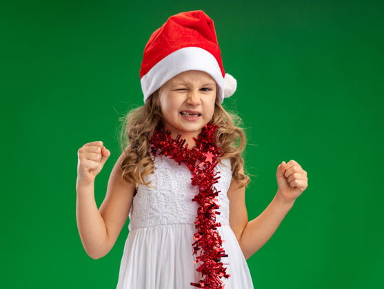圣诞节兴奋地眨着眼睛的小女孩戴着圣诞帽 脖子上戴着花环 在绿色的墙上显示“是”的手势小穿着展示