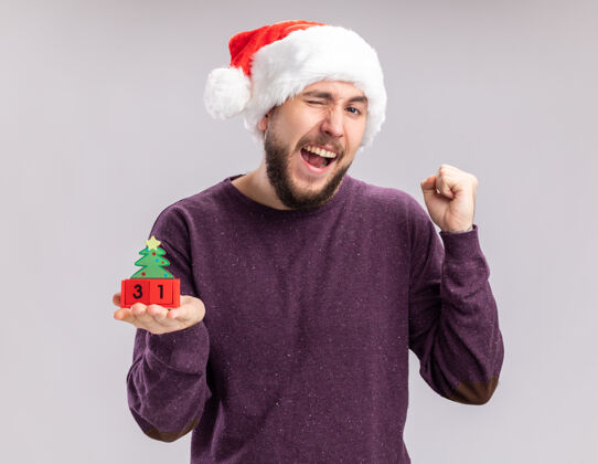 毛衣身穿紫色毛衣 头戴圣诞帽的年轻人站在白色背景上 展示着握紧新年拳头的方块帽子站着立方体