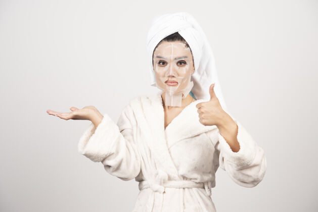 年轻女人竖起大拇指 穿着浴衣和带面罩的毛巾清洁人美容