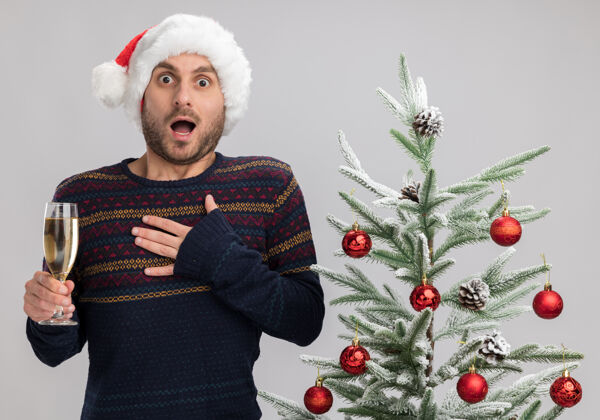 男人令人印象深刻的年轻白种人戴着圣诞帽站在圣诞树旁 手里拿着一杯香槟 手放在胸前 隔离在白墙上年轻人胸膛杯子