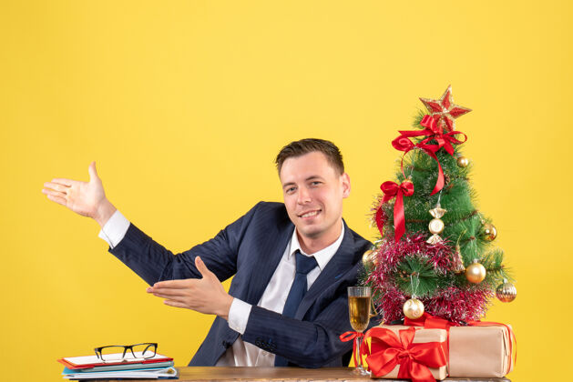男性正面图是一个快乐的年轻人指着坐在圣诞树旁桌子上的东西和黄色的礼物行政人员男人工作