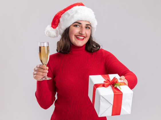 年轻人穿着红色毛衣 戴着圣诞帽 拿着一杯香槟和礼物的快乐女孩站在白色背景下 看着镜头 开心地微笑着站着礼物杯子