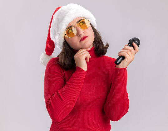 年轻人穿着红色毛衣 戴着圣诞帽 戴着眼镜 拿着麦克风 站在白色背景下 困惑地抬头看站着困惑圣诞老人
