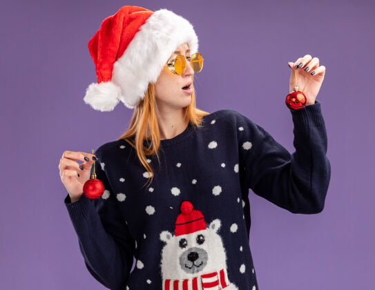 眼镜年轻漂亮的女孩穿着圣诞毛衣 戴着圣诞帽 戴着眼镜 拿着圣诞球看着紫色背景上的孤立看毛衣未租赁