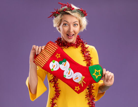 圣诞节兴奋的金发女郎头戴圣诞花环 脖子上戴着金箔花环 手里拿着圣诞长袜 看上去孤立地站在紫色的墙上头花环长袜