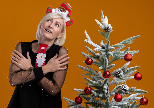 近令人印象深刻的中年金发女人戴着圣诞老人的头带和领带站在装饰圣诞树附近 双手交叉在手臂上 孤立地看着橙色的墙上女人圣诞节头带