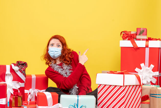 年轻黄色墙壁上戴着面具的年轻女子围坐在圣诞礼物旁的正视图Covid年轻女性病毒