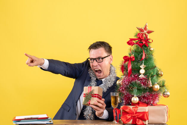 坐着愤怒的男人戴着眼镜坐在圣诞树旁的桌子前 黄色的墙上摆着礼物人人微笑