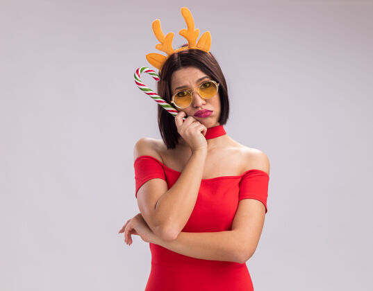 快乐无聊的年轻女孩戴着驯鹿鹿角头带和眼镜拿着圣诞糖果手杖用手抚摸着脸下巴圣诞快乐年轻的