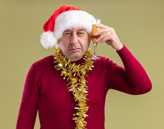 中间中年男子戴着圣诞老人的帽子 脖子上戴着金属丝 手里拿着一杯香槟 站在绿色的背景下 带着悲伤的表情看着相机圣诞节围着圣诞老人