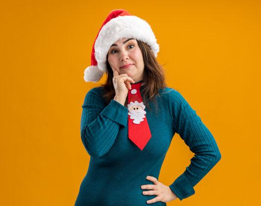 帽子戴着圣诞帽 系着圣诞领带的成年白种女人把手放在下巴上 看着隔离在橙色背景上的相机 还有复制空间印象深刻成人领带