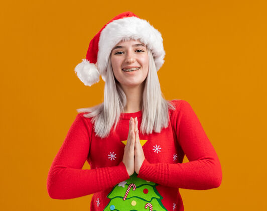 帽子穿着圣诞毛衣 戴着圣诞帽的金发女郎看着相机 双手合十 像纳马斯特一样 站在橙色的背景下 快乐而积极地站着站着积极年轻