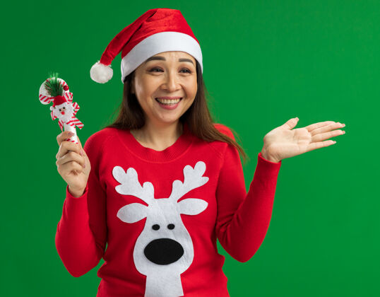 微笑快乐的年轻女子戴着圣诞老人帽 穿着红色毛衣 手持圣诞糖果手杖 脸上挂着微笑 站在绿色的背景下 望向一边圣诞节毛衣脸