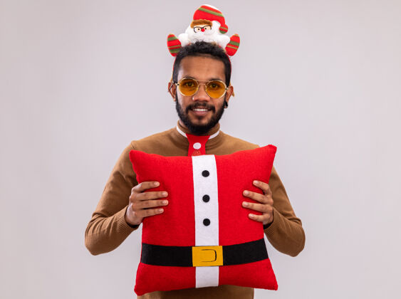 枕头一个非洲裔美国人 穿着棕色毛衣 头戴圣诞老人圈 打着有趣的红色领带 抱着圣诞枕头 站在白色背景下 面带微笑地看着摄像机站着领带抱着