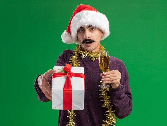 香槟年轻的留着胡子的男人戴着圣诞老人帽子 脖子上戴着金属丝 手里拿着一杯香槟和圣诞礼物 站在绿色背景下惊讶地看着摄像机惊讶圣诞节脖子