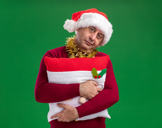 中间中年男子戴着圣诞老人帽 脖子上戴着金属丝 抱着圣诞枕头 站在绿色的背景下 带着悲伤的表情看着相机站着围着枕头