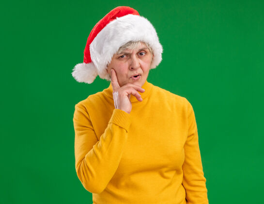 帽子带着圣诞老人帽子的体贴的老妇人把手放在下巴上 看着隔离在紫色背景上的相机 还有复印空间手体贴圣诞老人