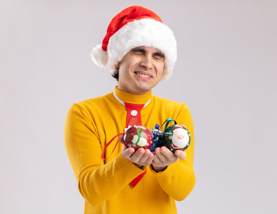 圣诞老人身穿黄色高领毛衣 戴圣诞帽 手持圣诞球的快乐年轻人站在白色背景上 欢快地微笑着站着微笑帽子