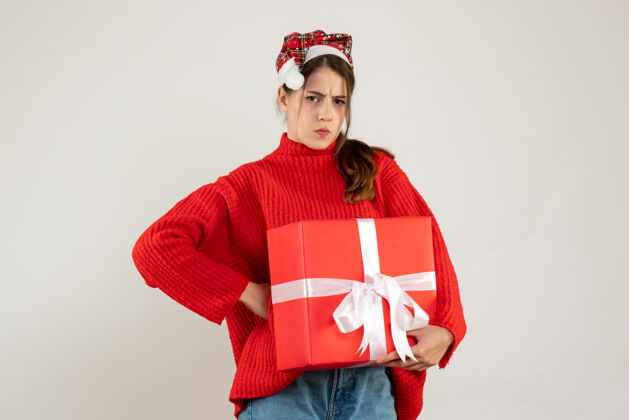 长袍不满意的女孩戴着圣诞帽拿着礼物手放在腰上站在白色的圣诞老人和服站着