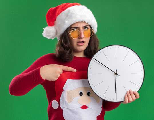 站着穿着圣诞毛衣戴着圣诞帽戴着眼镜拿着挂钟用食指指着它看着相机严肃的脸站在绿色的背景上手指圣诞老人眼镜