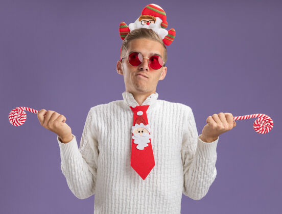 头带迷茫的年轻帅哥戴着圣诞老人的头带和领带戴着眼镜拿着圣诞糖果手杖看着隔离在紫色墙上的一面手杖领带年轻