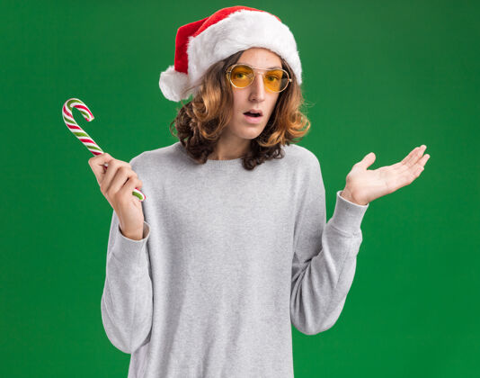 手杖戴着圣诞老人帽 戴着黄色眼镜 拿着糖果手杖的年轻人看着摄像机 困惑地微笑着 举起双臂站在绿色的背景上圣诞老人眼镜帽子